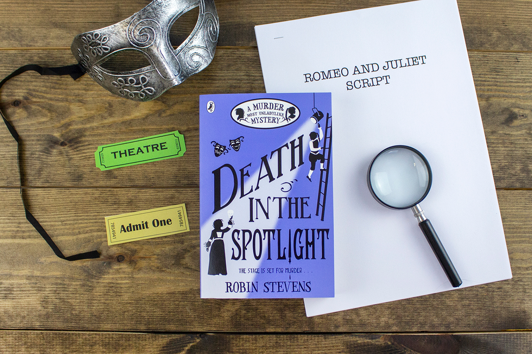 A image of Robin's Steven's book Death in the Spotlight on a wooden table background surrounded by theatre tickets, a Romeo & Juliet script, a silver masquerade mask and a magnifying glass