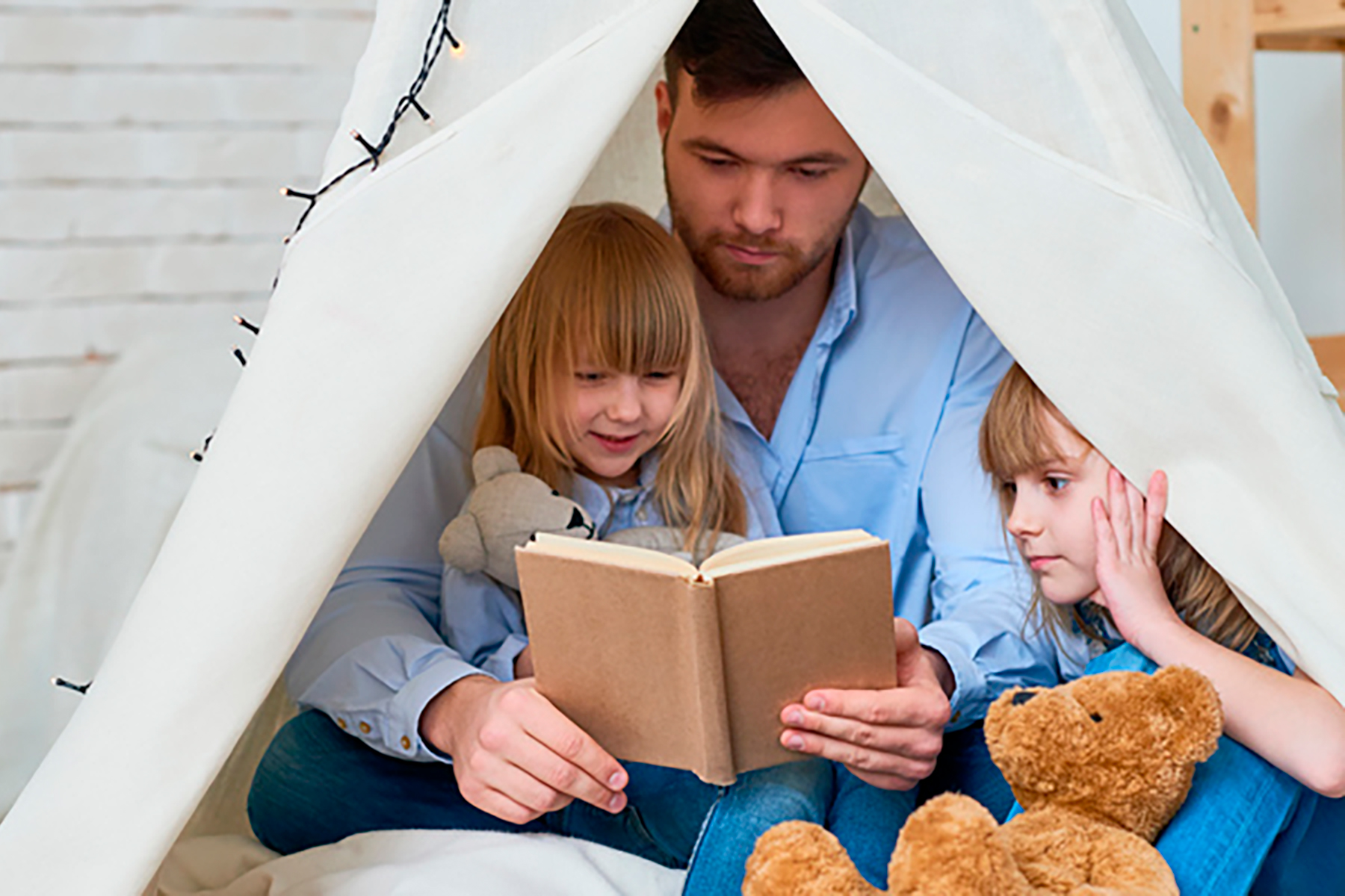 A photo of a dad sitting under an indoor canopy with his two young girls as he reads a story to them