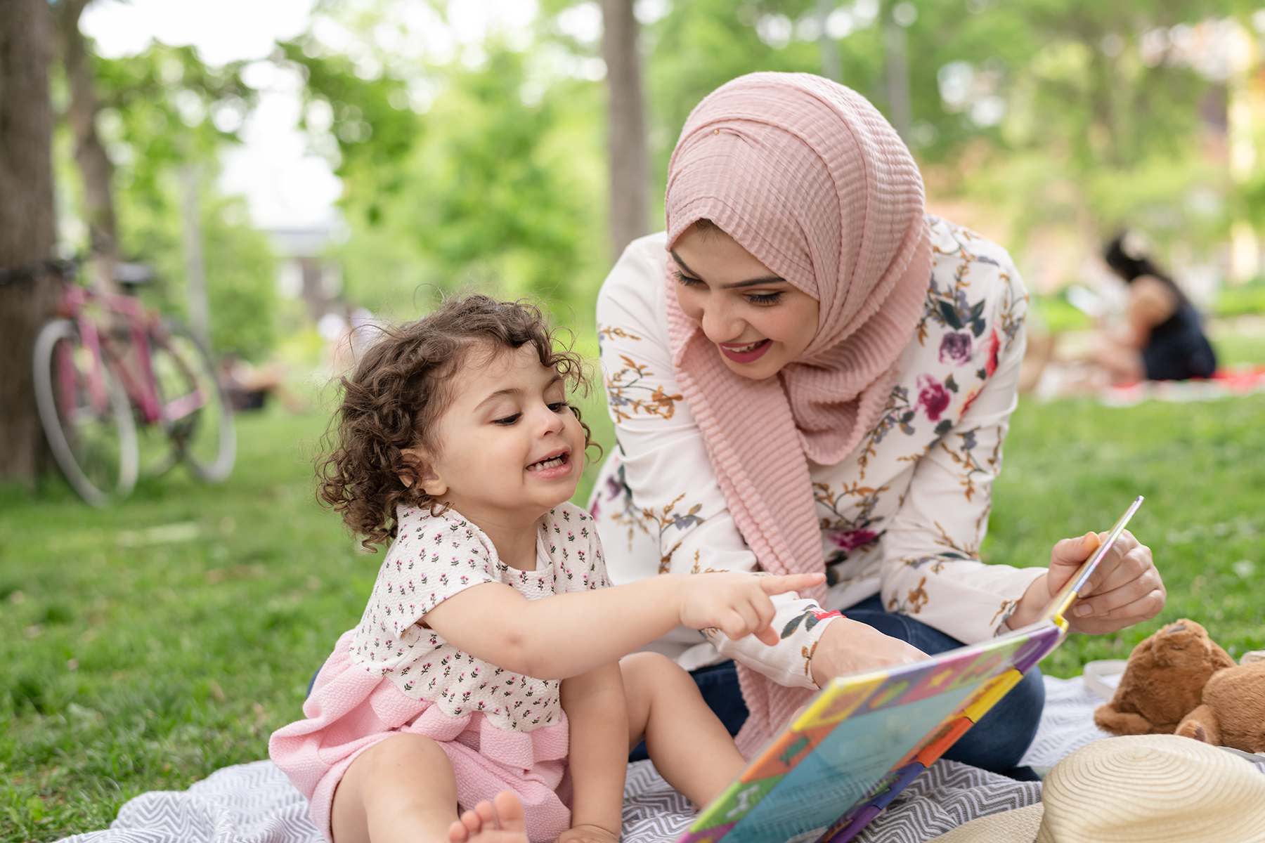 A photo of a mother sitting in a park with her young child. They are reading and interacting with a book