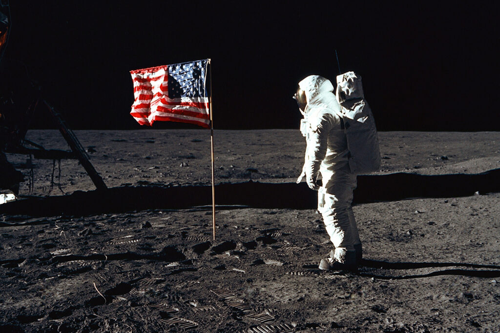 A photo by NASA of astronaut Buzz Aldrin on the moon. He is facing an American flag that as been stuck in the surface of the moon