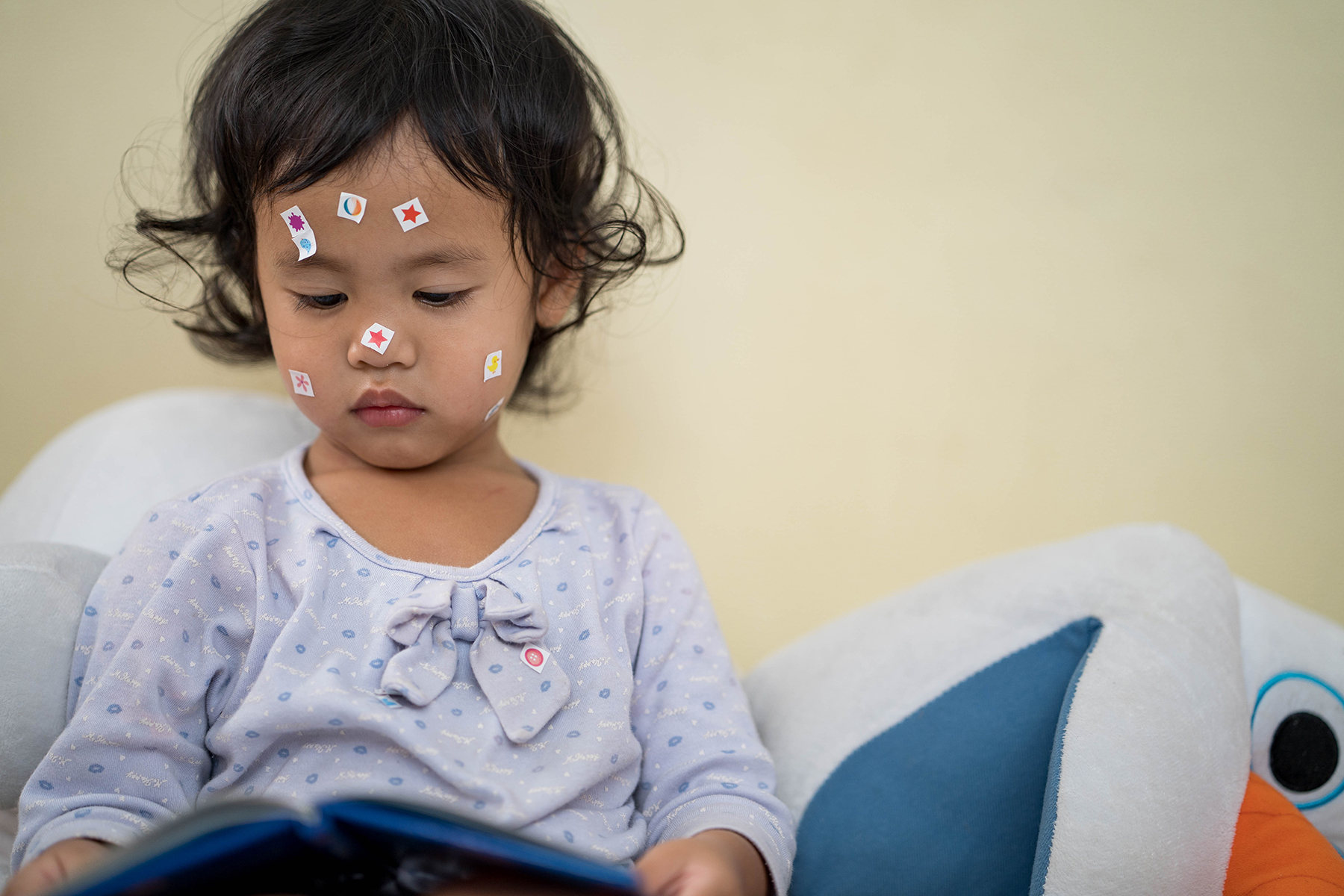 Photo of a little girl sitting on her bed, looking at a book with some stickers on her face
