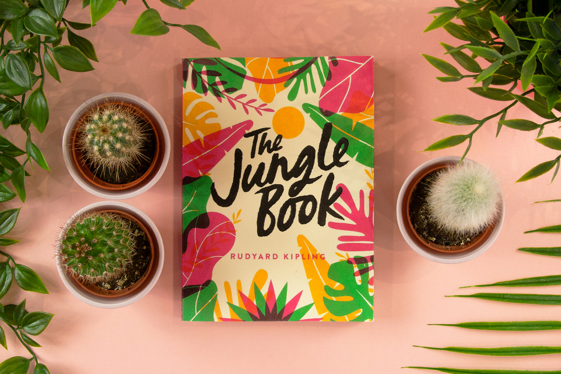 A photo of the book The Jungle Book by Rudyard Kipling on a light peach background surrounded by cacti and plants