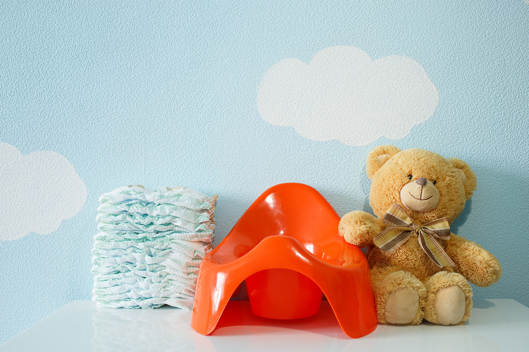 A photo of an orange potty next to a teddy bear and nappies. The background is a light blue wall with white clouds