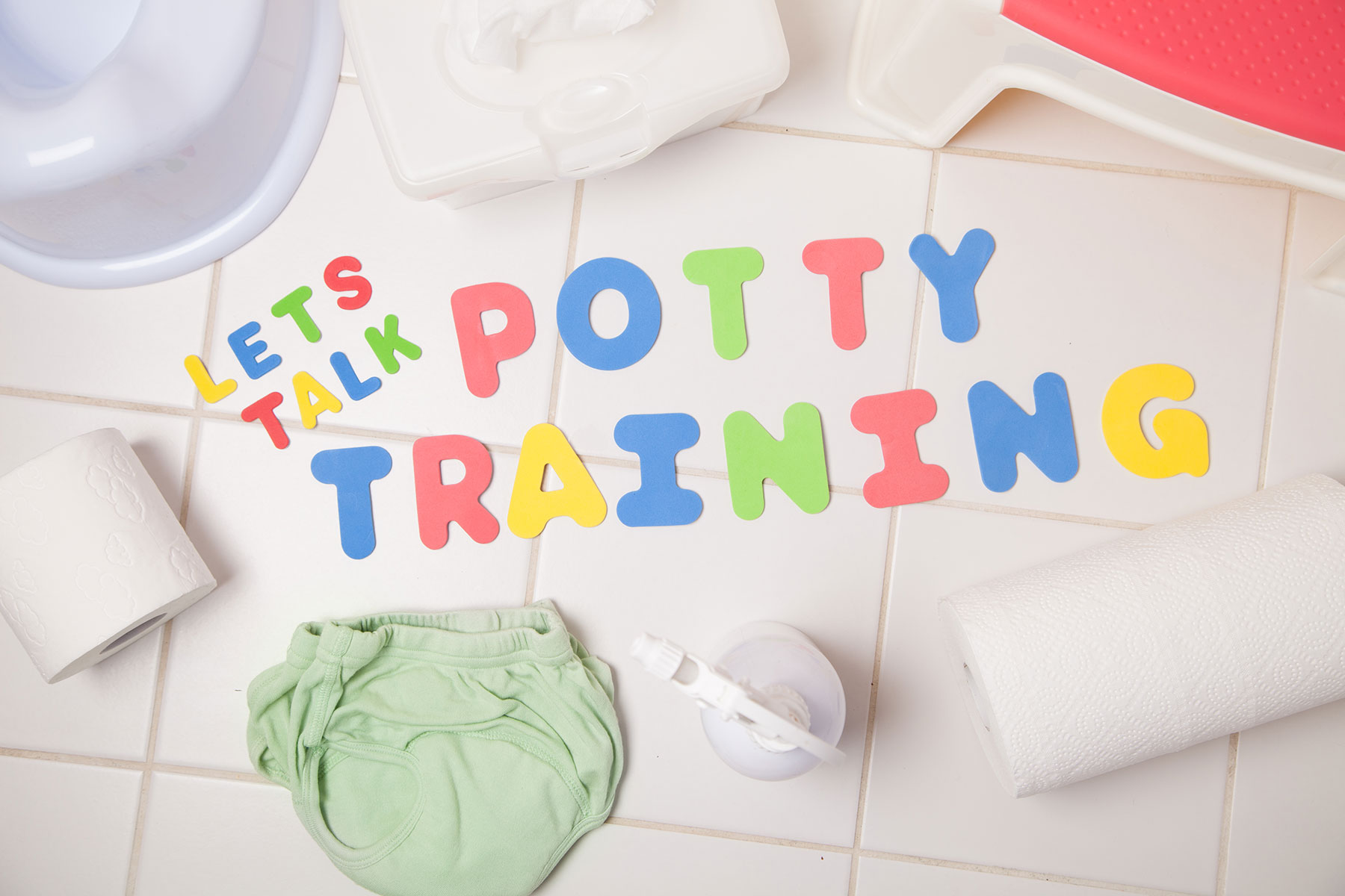 A photo of the words 'Let's Talk Potty Training' on the floor of a white bathroom surrounded by toilet paper, wipes, green pants and a potty