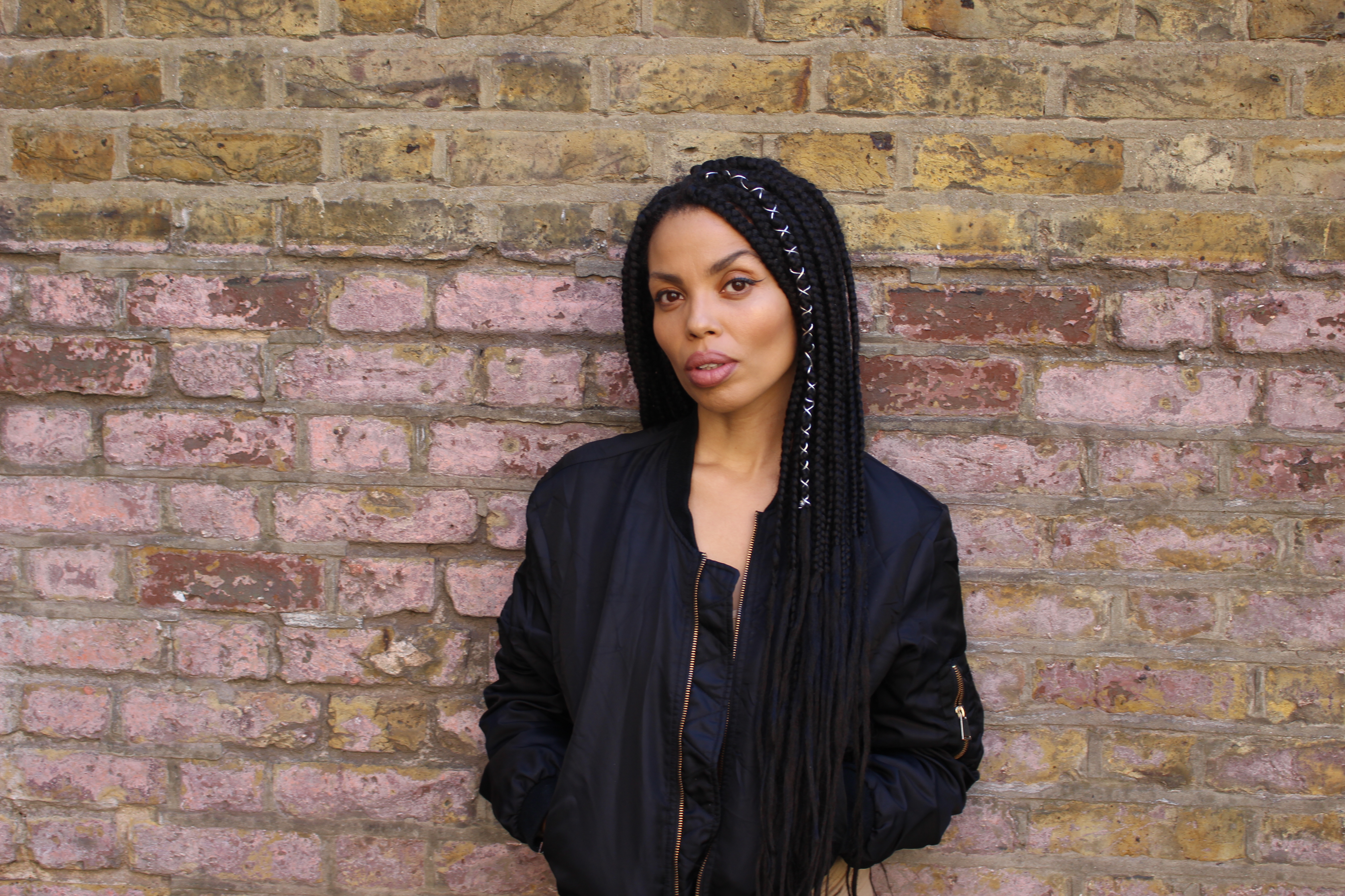 Photo of Emma Dabiri wearing a black top and black jacket, standing in front of a brick wall.