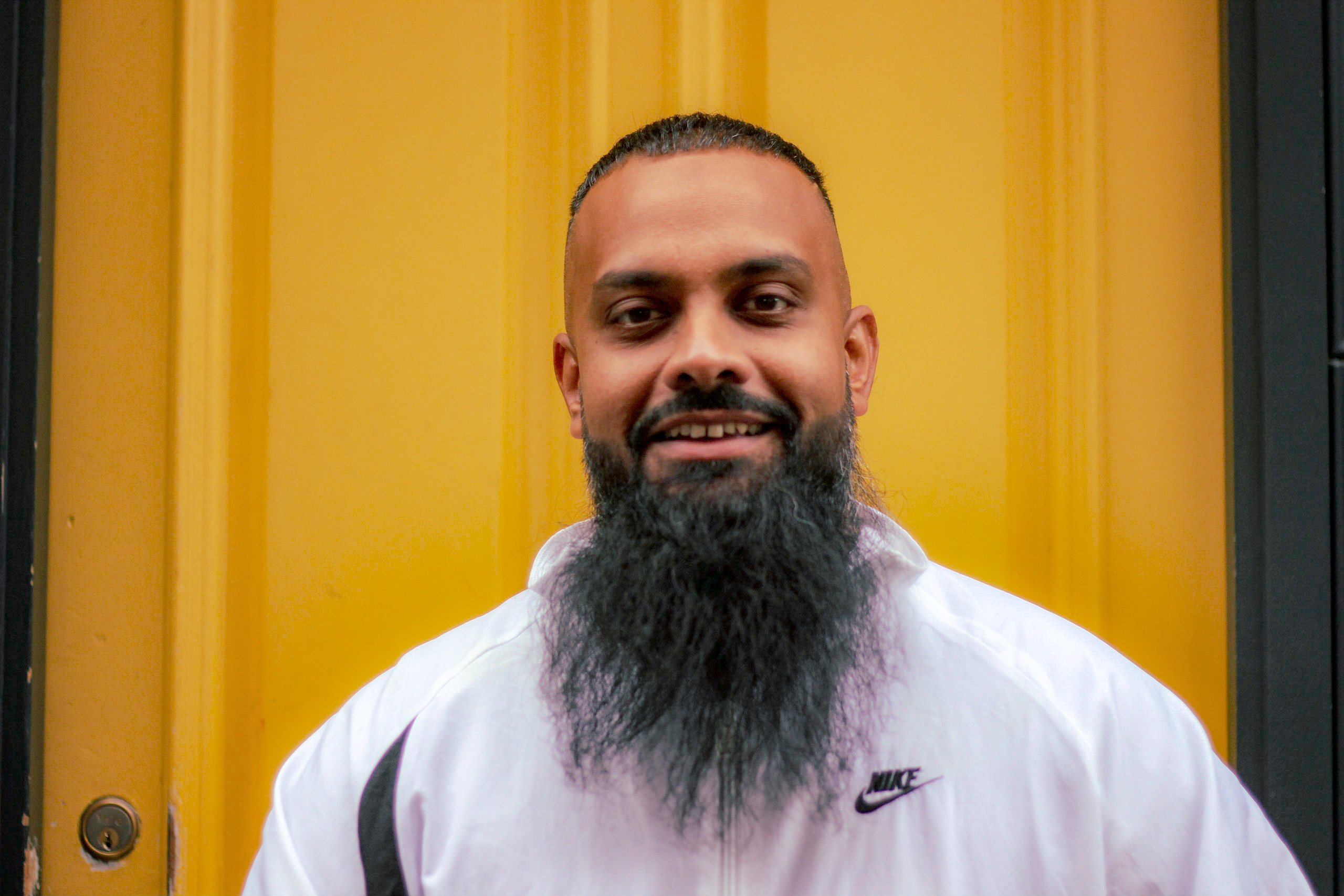 Picture of Guz Khan wearing a white Nike jacket, standing against a yellow background.