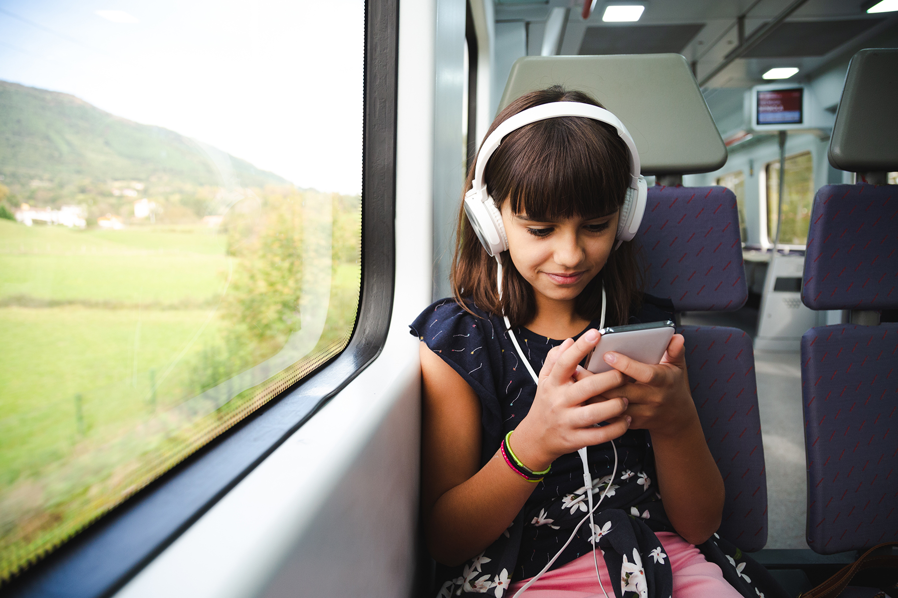 A photo of a young girl sitting on a train - she has white headphones on and is looking at her phone whilst she plays an audiobook