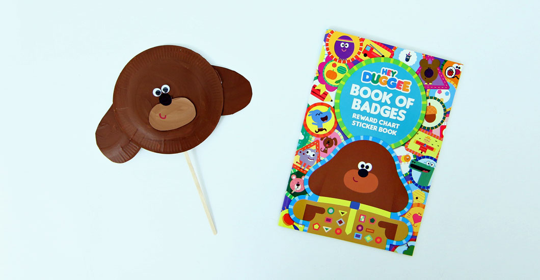 A photo of a homemade Hey Duggee mask on pale blue background next to the book Hey Duggee's Book of Badges