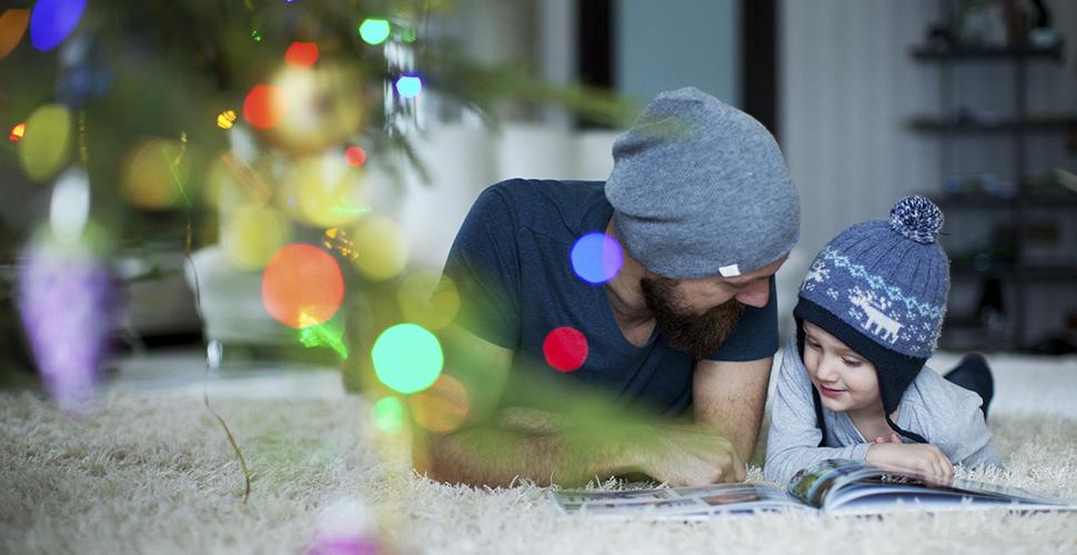 A photo of a dad and his son laying on the floor and reading a book with a Christmas tree in the background