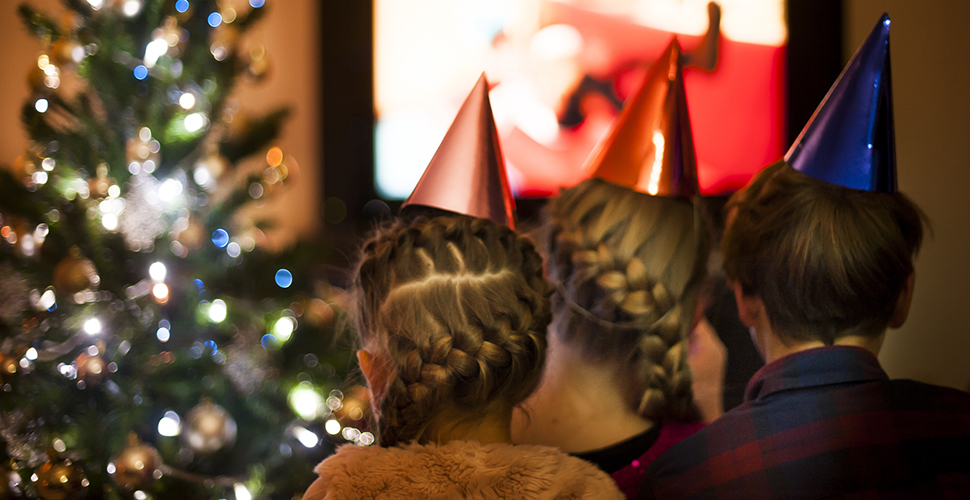 A photo of three children wearing party hats as they sit in front of a television screen with a Christmas tree in the background