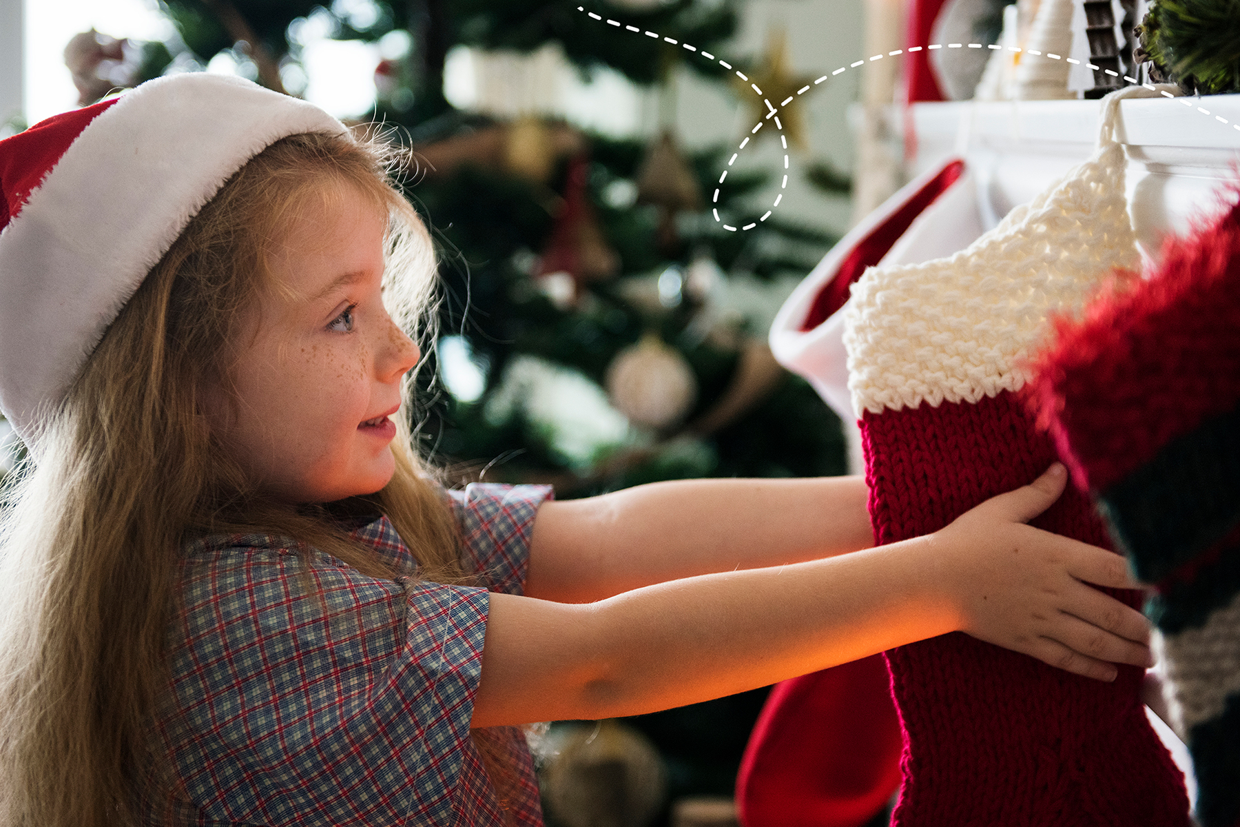 An photo of a little girl wearing a Santa hat and holding a stocking that is hooked onto a fireplace
