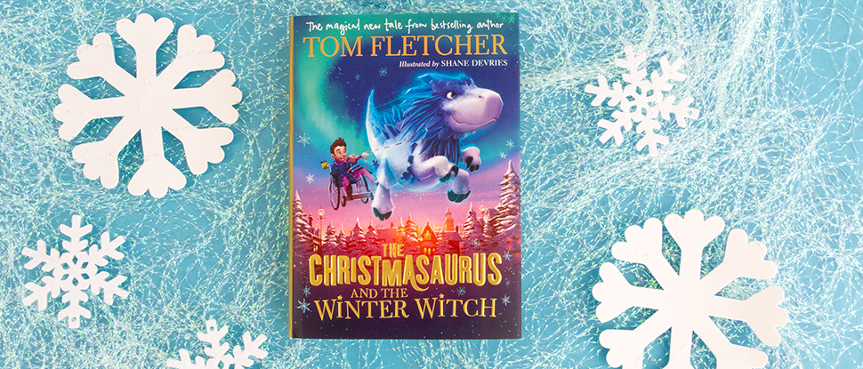 A photo of a copy of The Christmasaurus and the Winter Witch by Tom Fletcher on a light blue background alongside white paper snowflakes