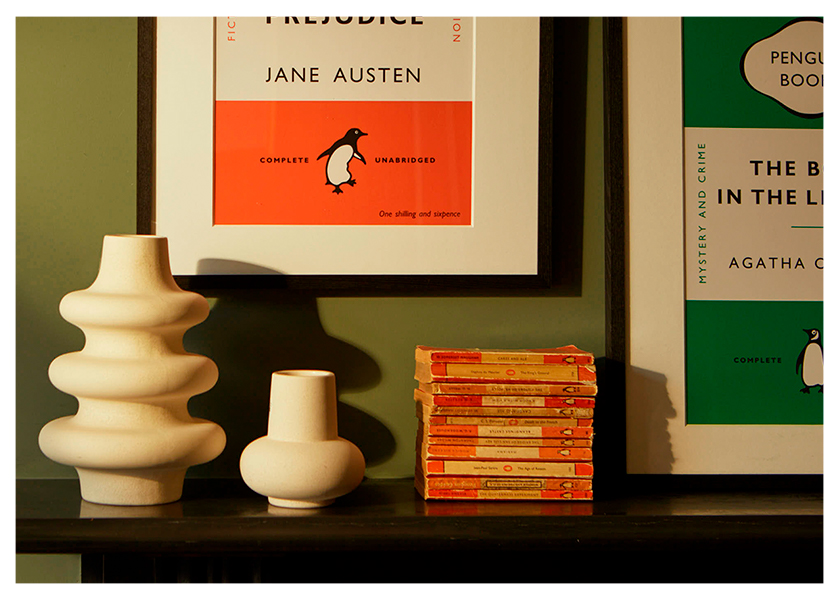 A wall showing the parts of two framed Penguin prints, with a shelf of classic Penguin novels and some vases.