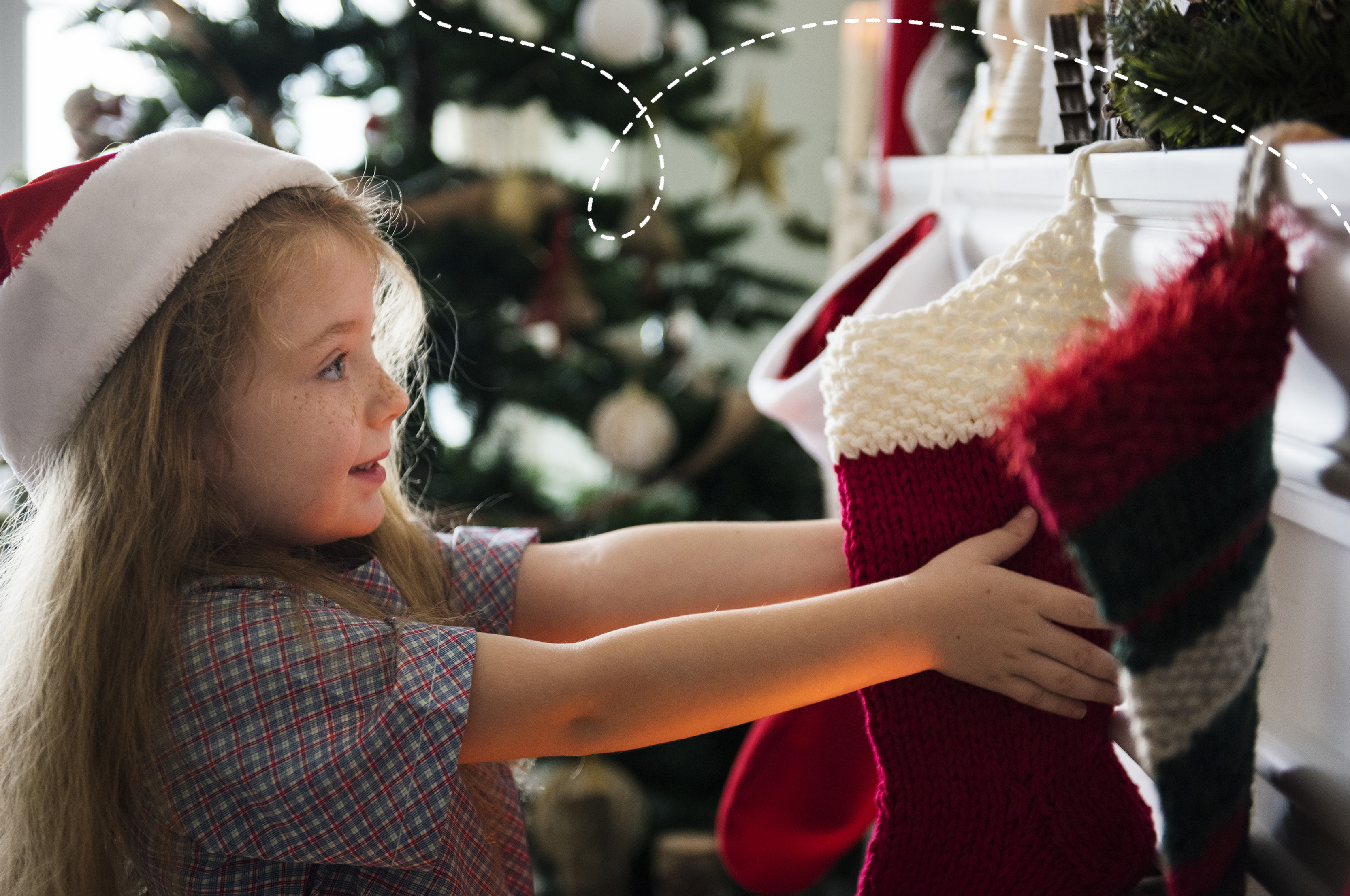 An photo of a little girl wearing a Santa hat and holding a stocking that is hooked onto a fireplace