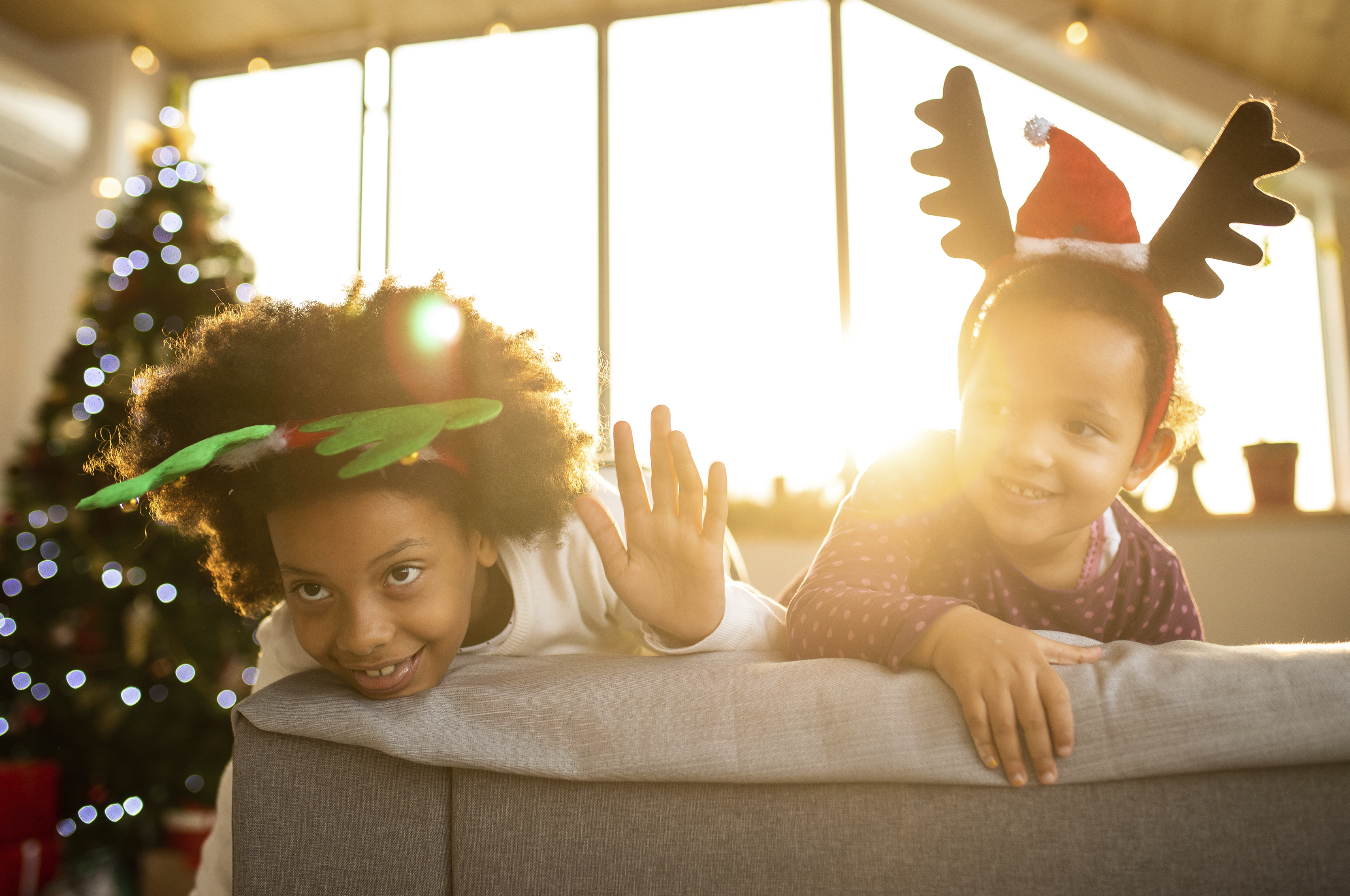A photo of a couple of children on a sofa wearing festive antlers with a Christmas tree in the background