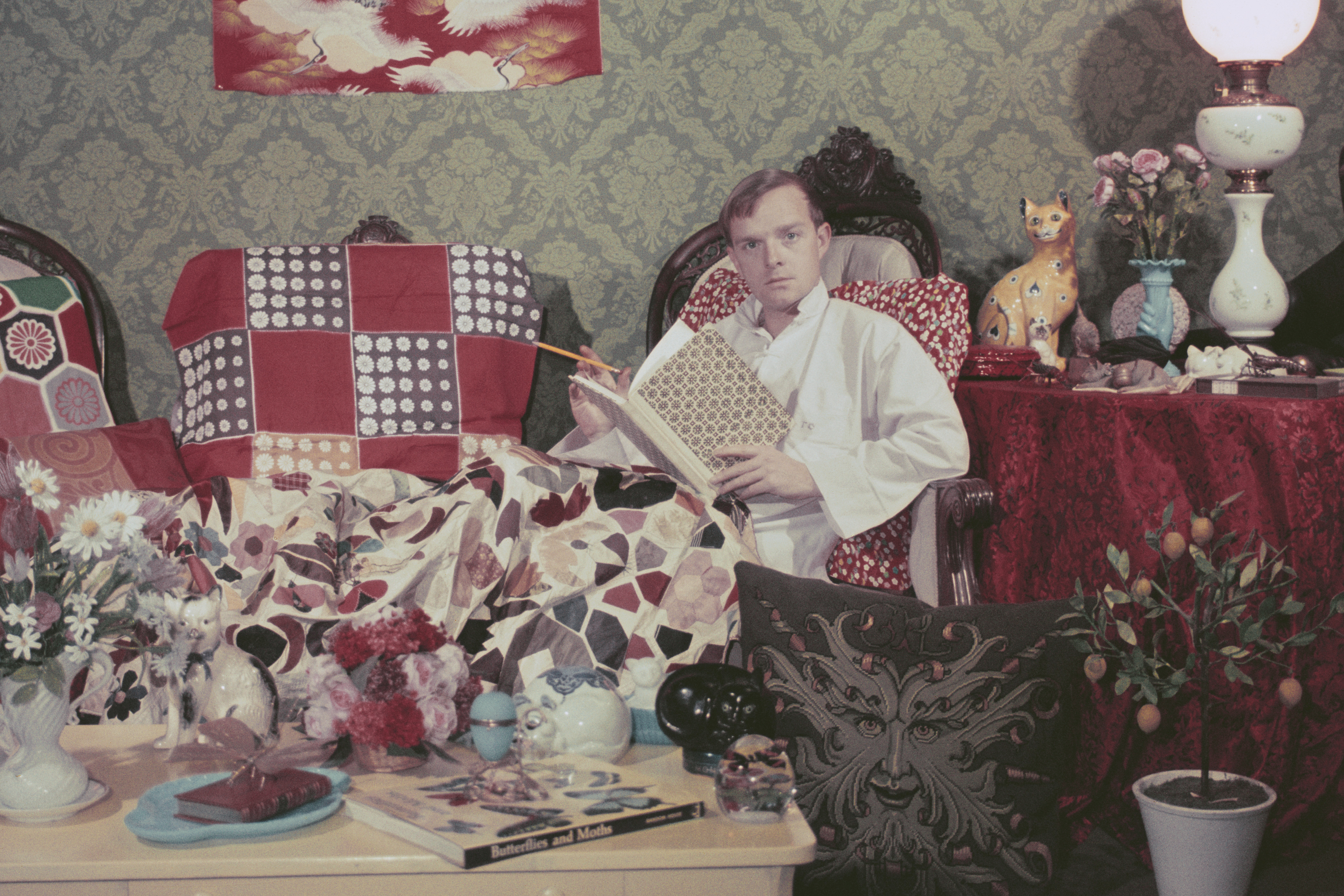 A photograph of Truman Capote, at home, sitting in a white robe with a notebook, surrounded by blankets and ornaments