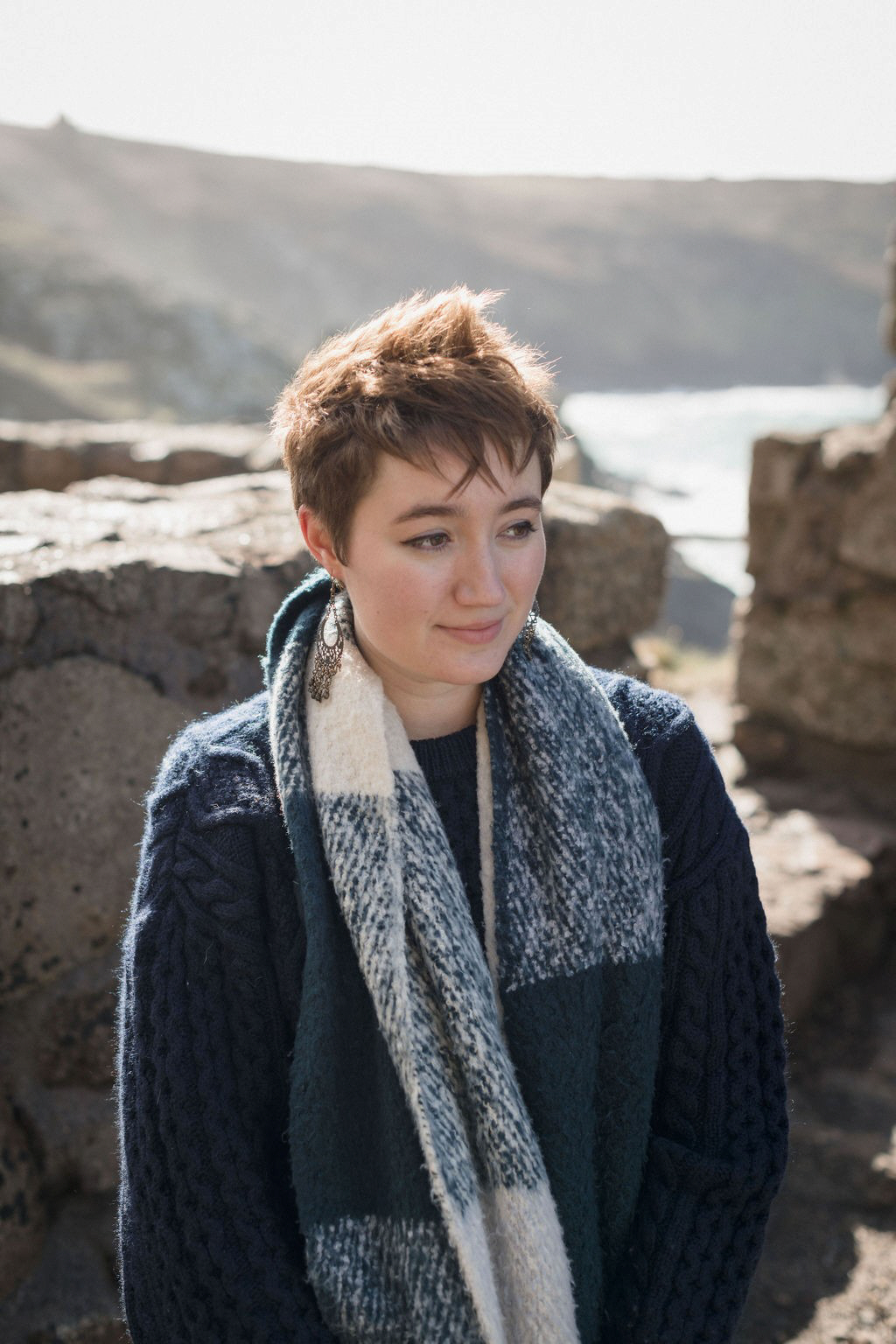 Photograph of Anna Bailey, wearing a dark jumper and blue and white striped scarf, in front of some rocks, looking off to the right.