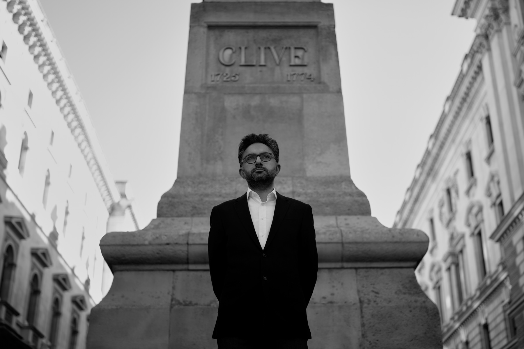 Black and white photograph of Sathnam Saghera, with his hands behind his back, in front of a statue of Sir Robert Clive in Whitehall.