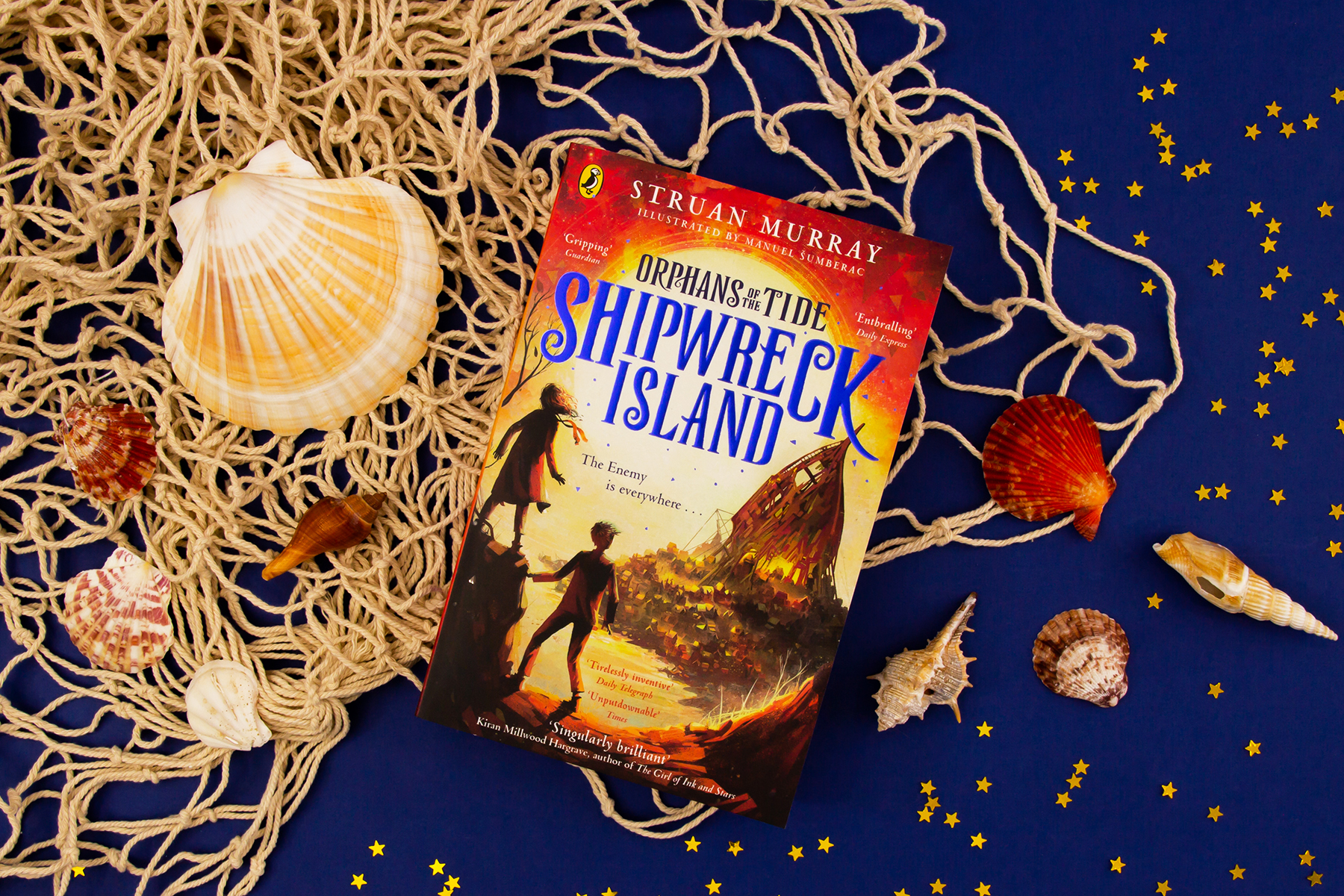 A photo of the book Shipwreck Island by Struan Murray on a dark blue background surrounded by a fishing net and shells