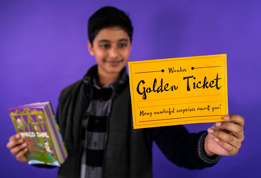 A photo of a young boy holding a copy of Charlie and the Chocolate Factory and a 'golden ticket' from the book. He is standing against a bright purple background