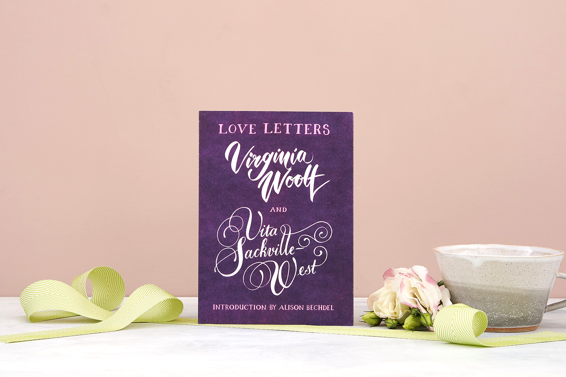 Lifestyle shot of Love Letters: Vita and Virginia, standing on a white marble base against a pale pink wall. A green fabric ribbon is draped across the front of the shot, and a mug and pink flowers stand to the left of the book.