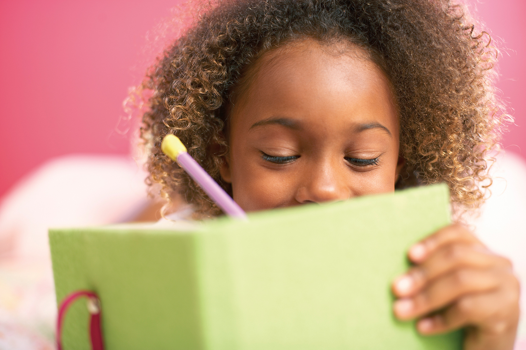A photo of a young girl writing in her green diary on a pink background