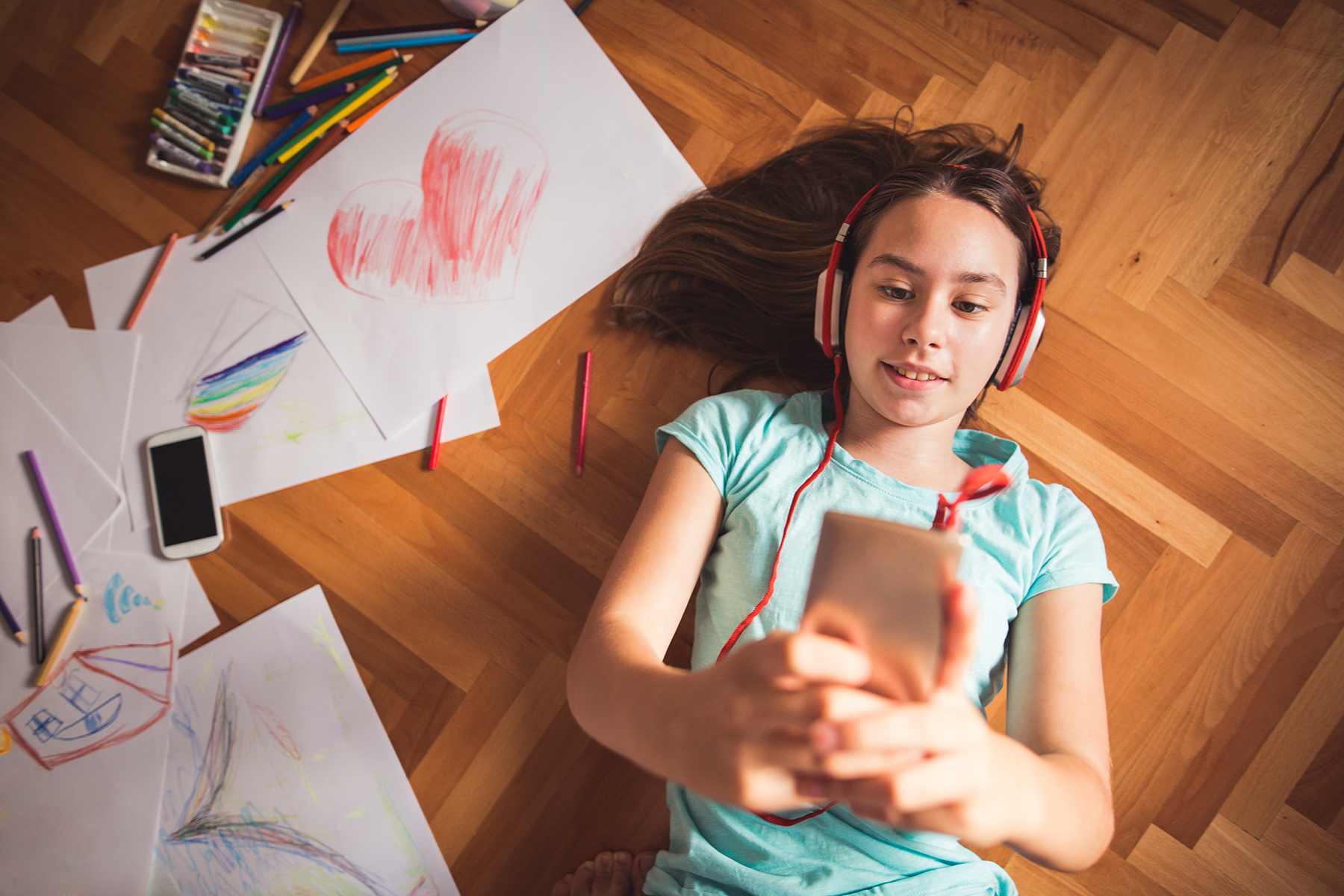 A photo of a young girl laying on the floor with a pair of headphones on, looking at her phone whilst she plays an audiobook