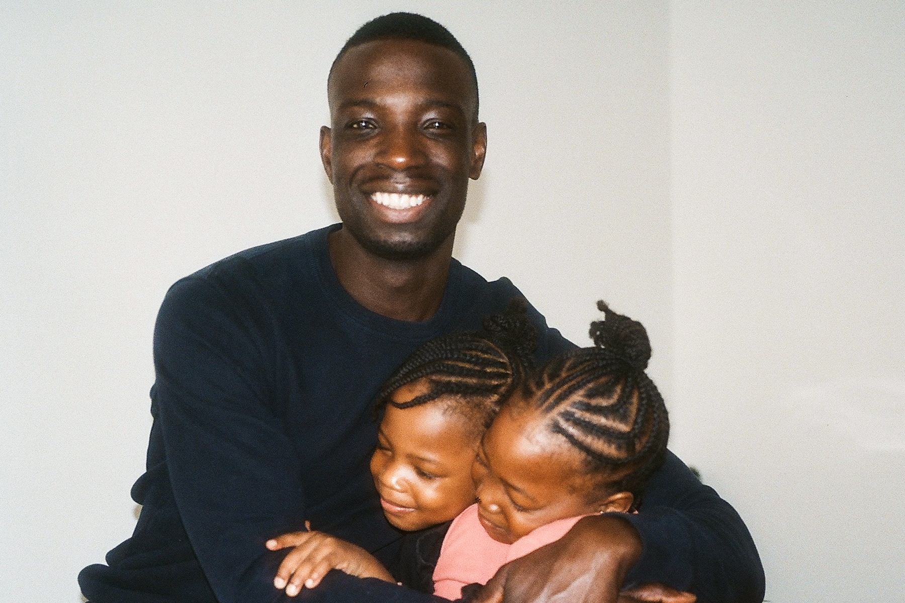 A photo of father Baff smiling and cuddling his two children against a white wall