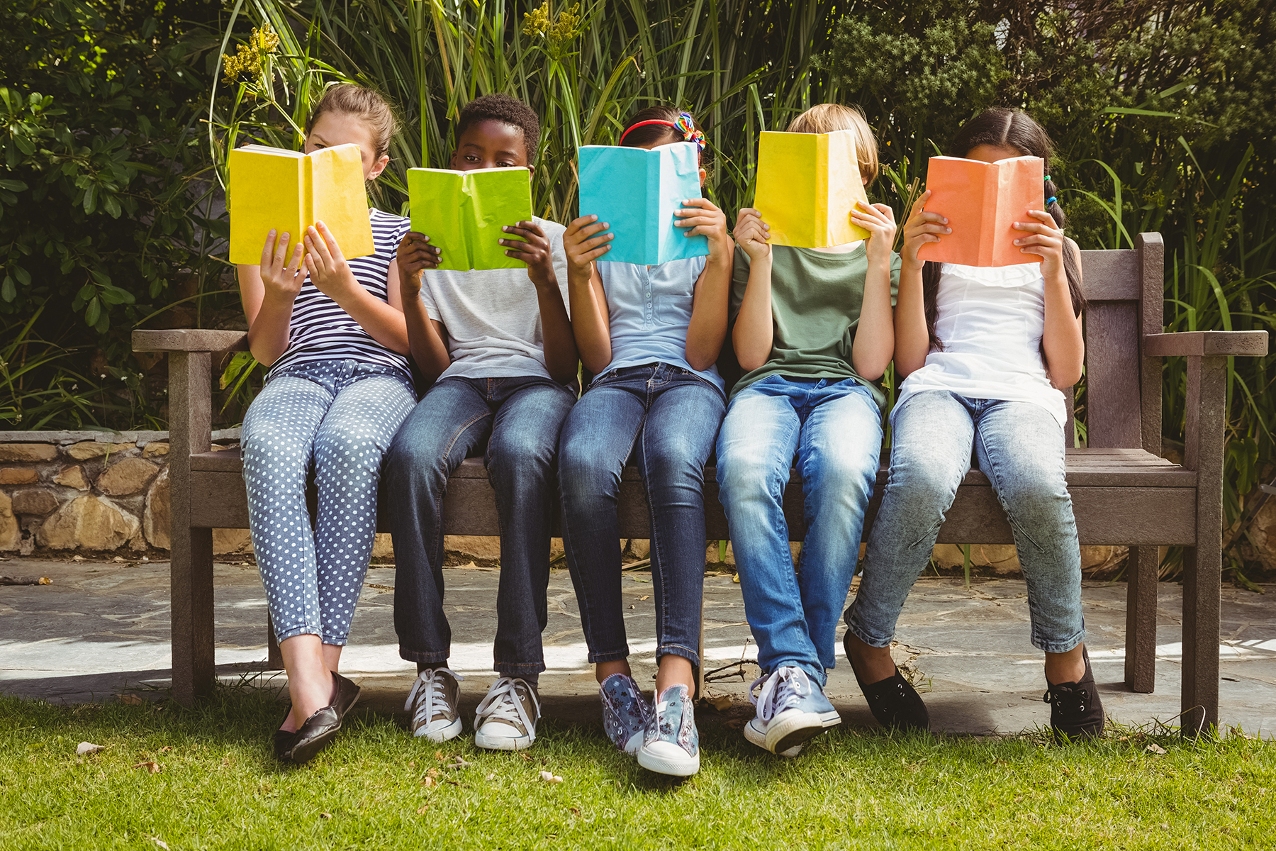 A photo of five children, a mixture of boys and girls, all sitting on a bench in the park, holding up books that cover their faces