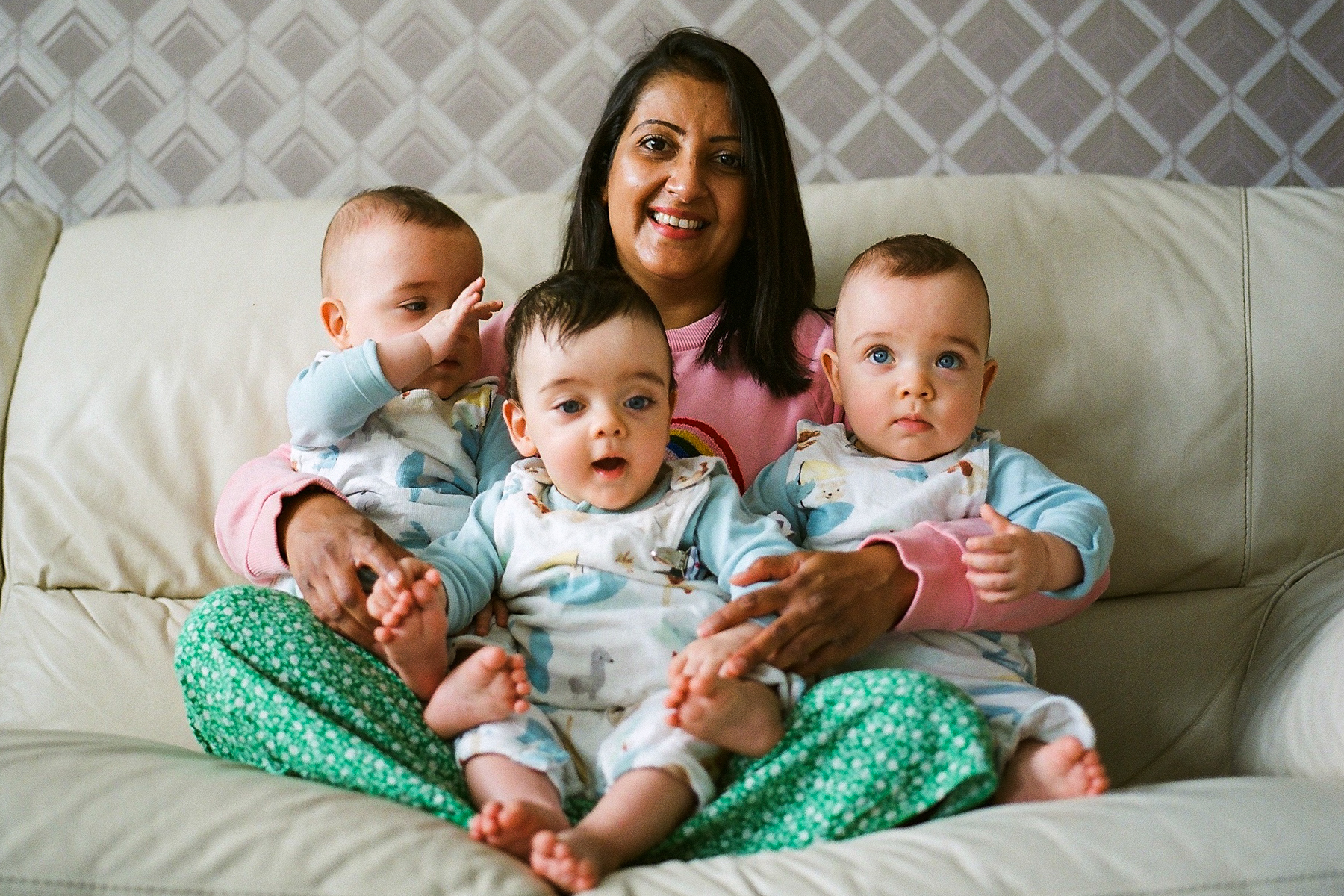 A photo of mum Kreena smiling into the camera and sitting on her bed with her triplets in her lap