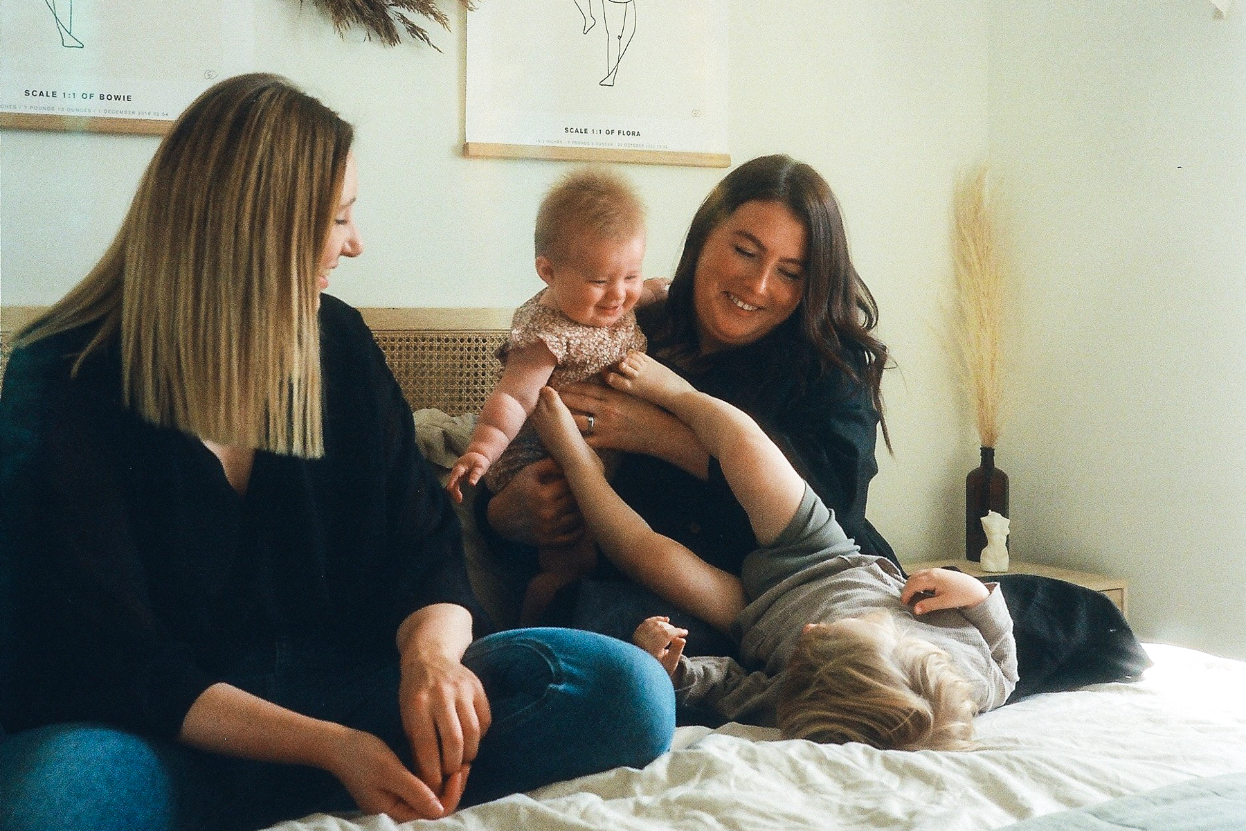 A photo of mums Lauren and Khiana with their two children; they are all sitting on a bed, Lauren is holding their youngest who is looking down and smiling at her older sibling who is laying on the bed with their feet up in the air