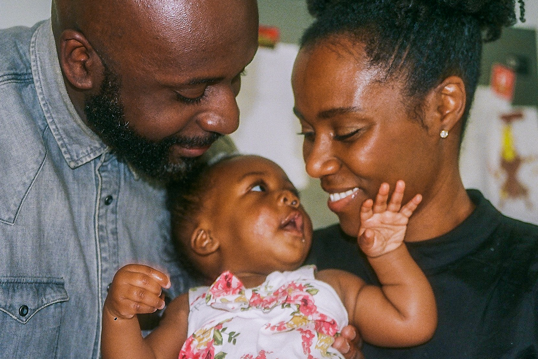 A close up photo of father Oke with his daughter and wife; Oke and his wife are both looking at their daughter whilst she is looking up at her mother