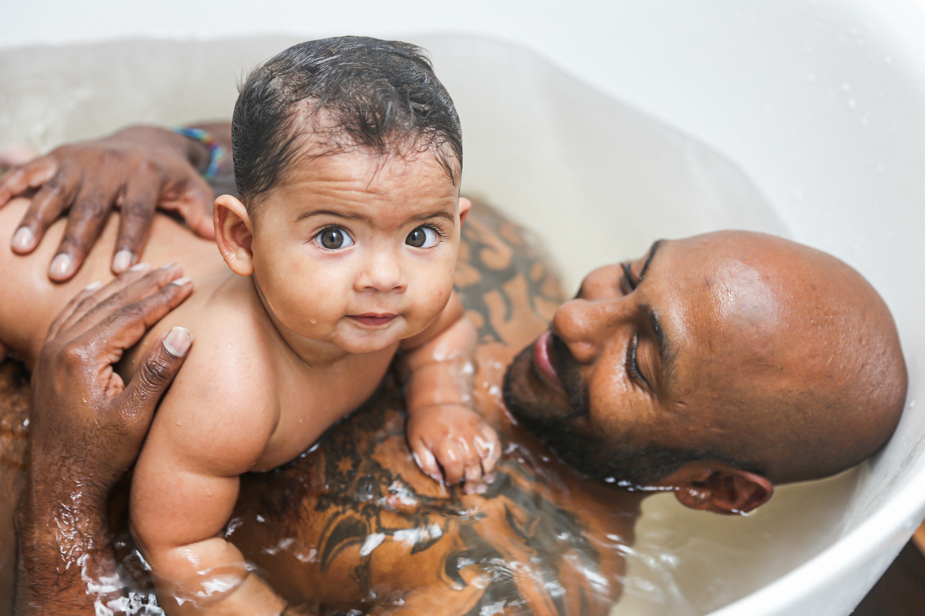 A photo of father Prema in the bath; he is laying down and holding his child on his chest who is looking directly into the camera