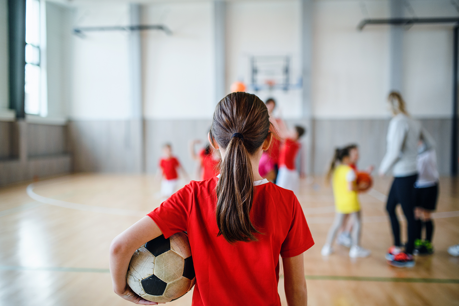 A photo of a young girl with her back to the camera, standing in a sports hall with a football under her arm. There are other children in the background