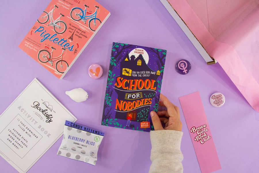 A photo of the contents of the Brave Girls Book Club's subscription box; there are a couple of books alongside an activity book, a bookmark, badges and sweet treats on a pale purple background