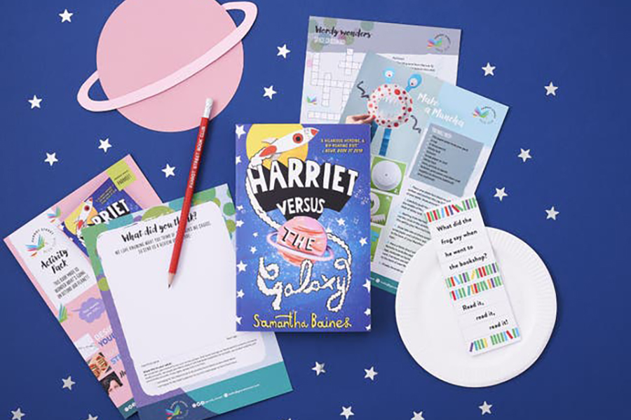 A photo of the contents of the Parrot Street Book Club's subscription box; there is a book alongside some activity sheets and a pencil on a dark blue background with some silver stars and a pink planet