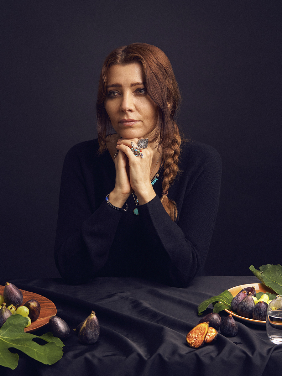 A photograph of Elif Shafak, dressed in black, against a black background, above a table with figs and fig leaves on it.