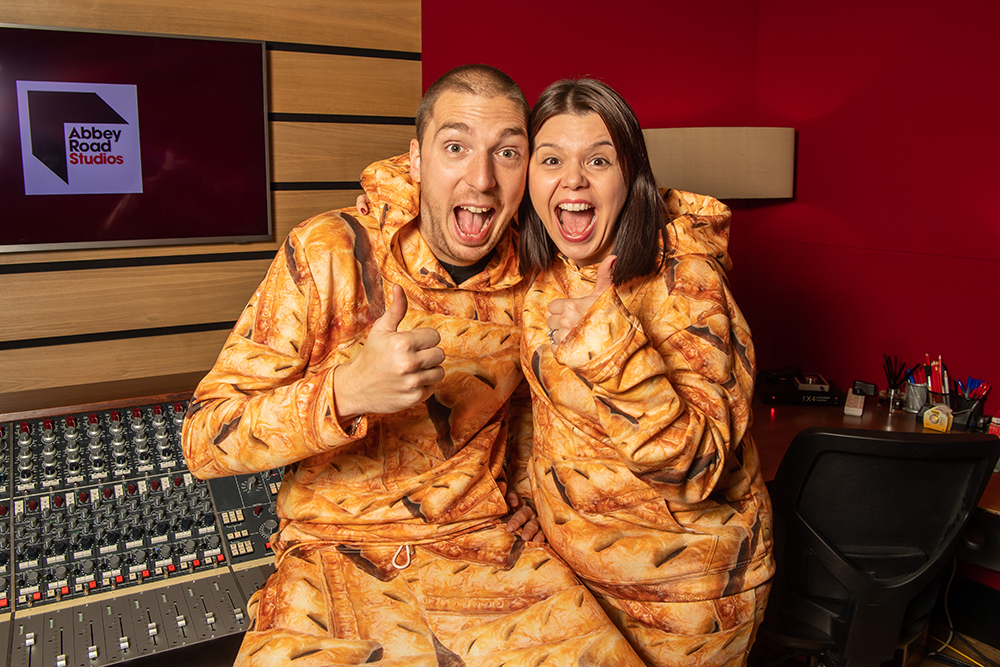 A photo of authors and LadBaby creators Mark and Roxanne Hoyle smiling/laughing whilst holding their thumbs up. They are both dressed in sausage roll print tracksuits and are in the Abbey Road Studio