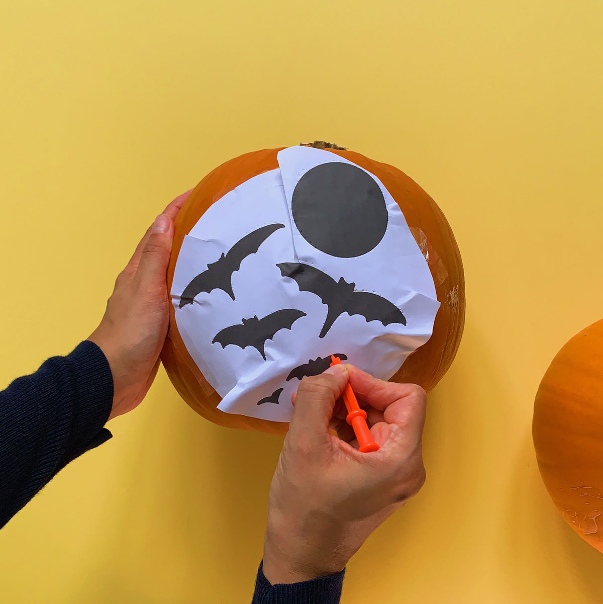 A photo of a stencil taped to a pumpkin and hands using a poker to poke the design out