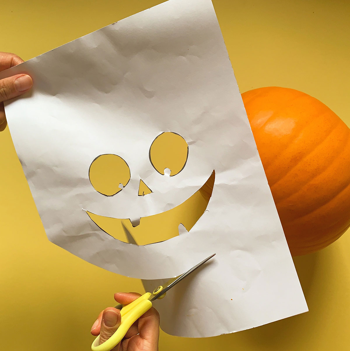 A photo of a pair of hands cutting out a pumpkin face stencil from paper with a pair of scissors and a pumpkin in the background