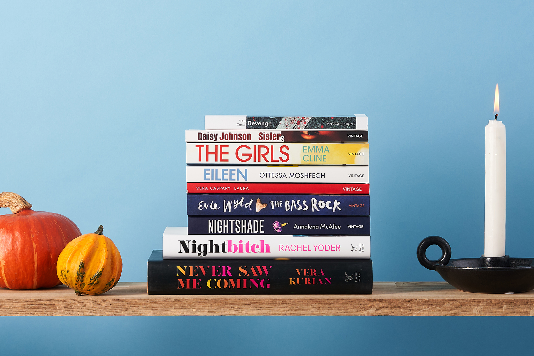 Stack of 9 books on a wooden shelf against a blue background, surrounded by gourds and a white candle.