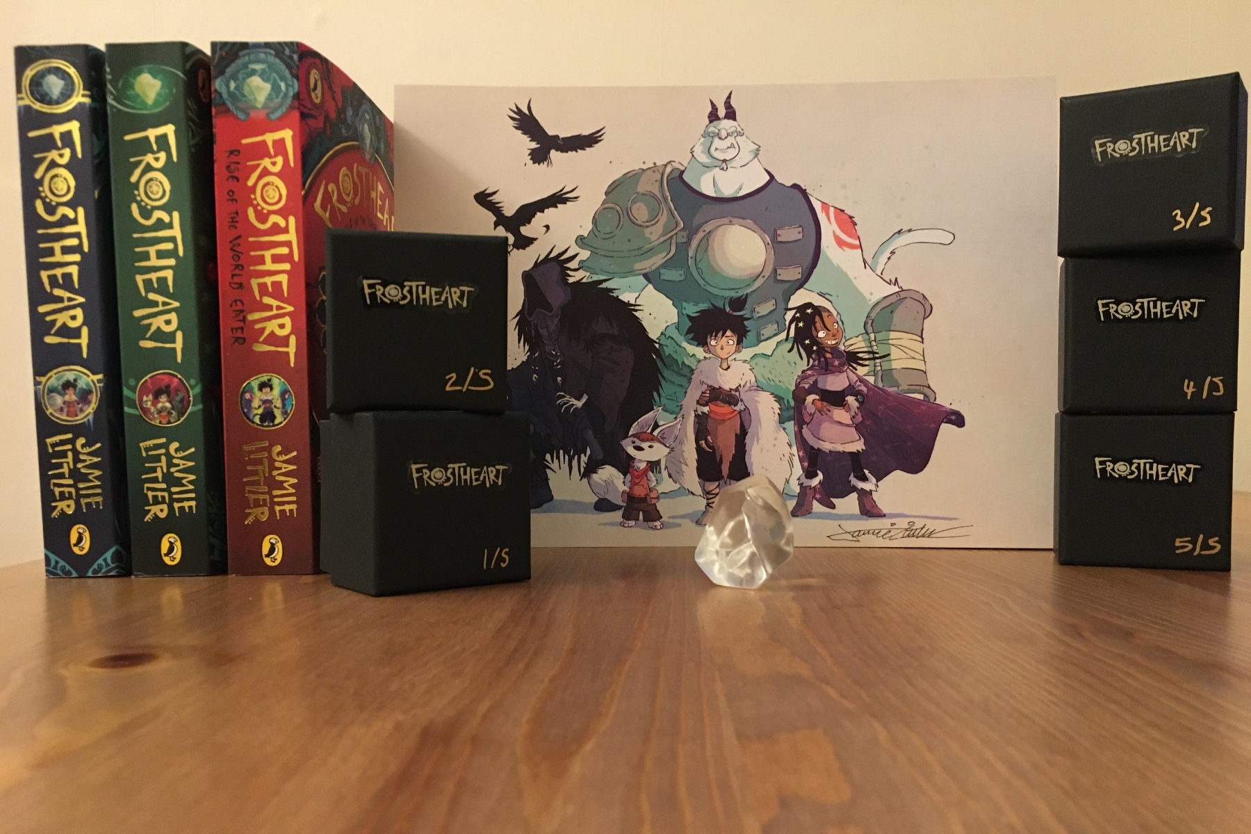 A photo showing a frost-heart stone, the three books in the series and a signed print on a wooden table