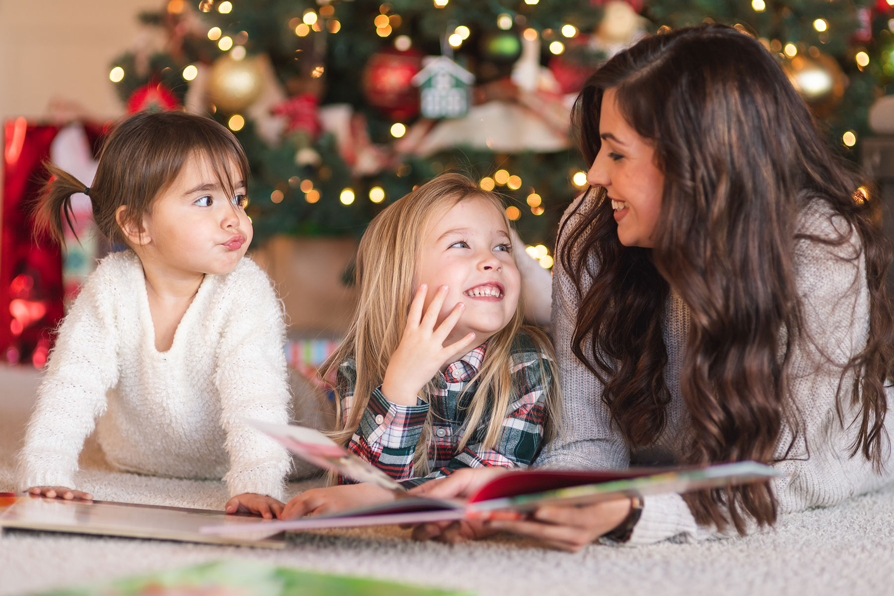 A photo of a mother laying on the floor with her two daughters and reading to them with a Christmas tree in the background. The two girls are looking at their mother who is smiling back at them