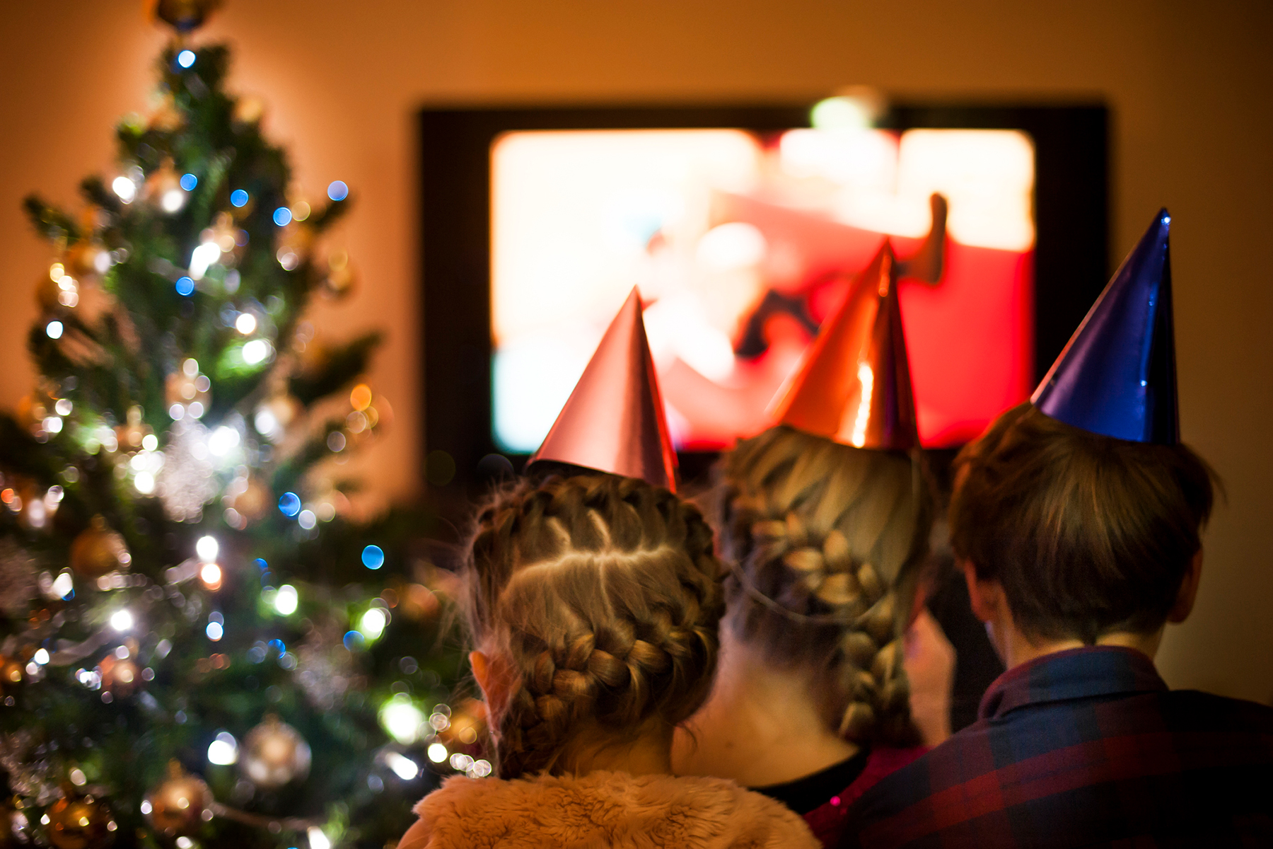 A photo of three children wearing party hats as they sit in front of a television screen with a Christmas tree in the background