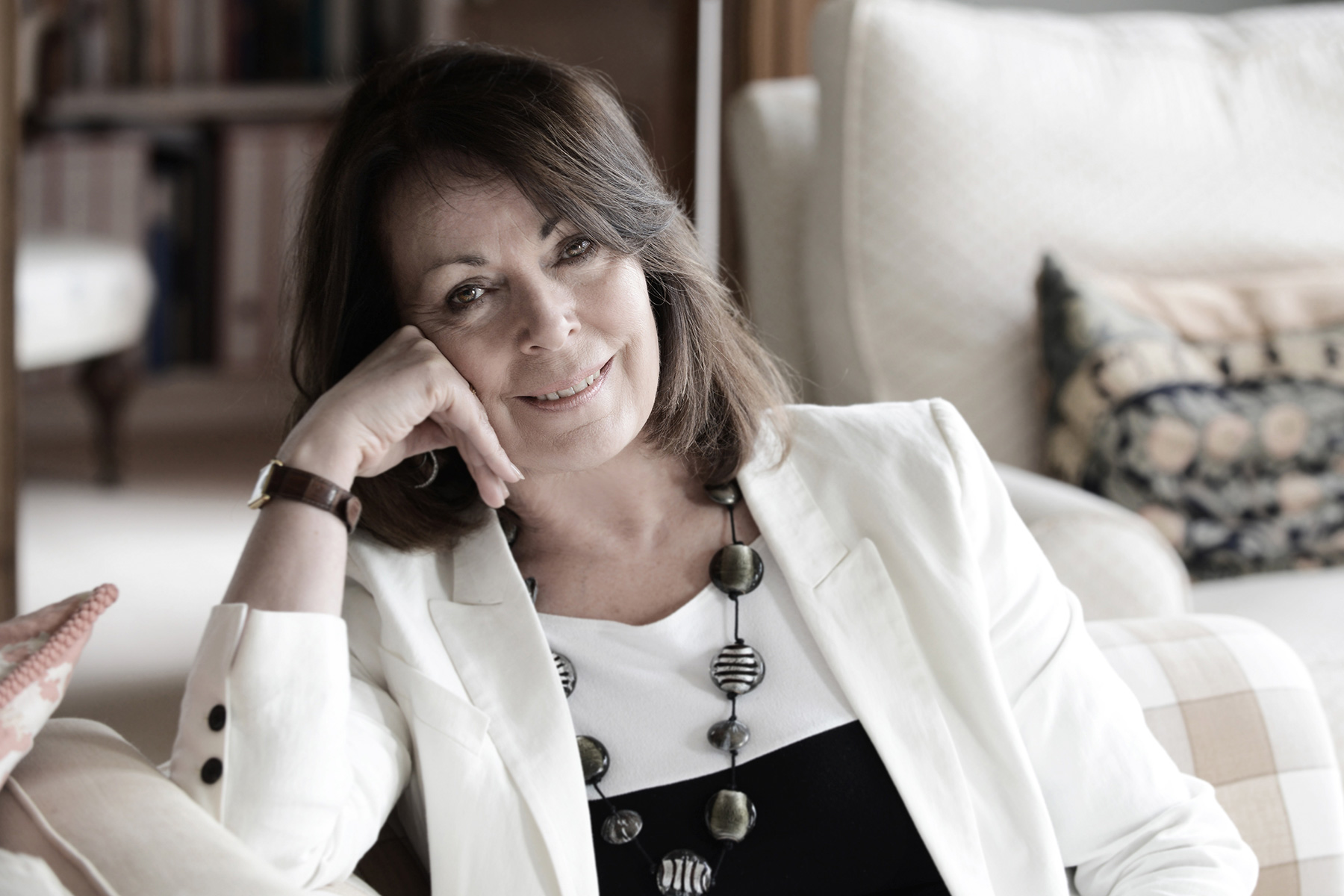A photograph of Rose Tremain sitting on a white sofa, wearing a white blazer