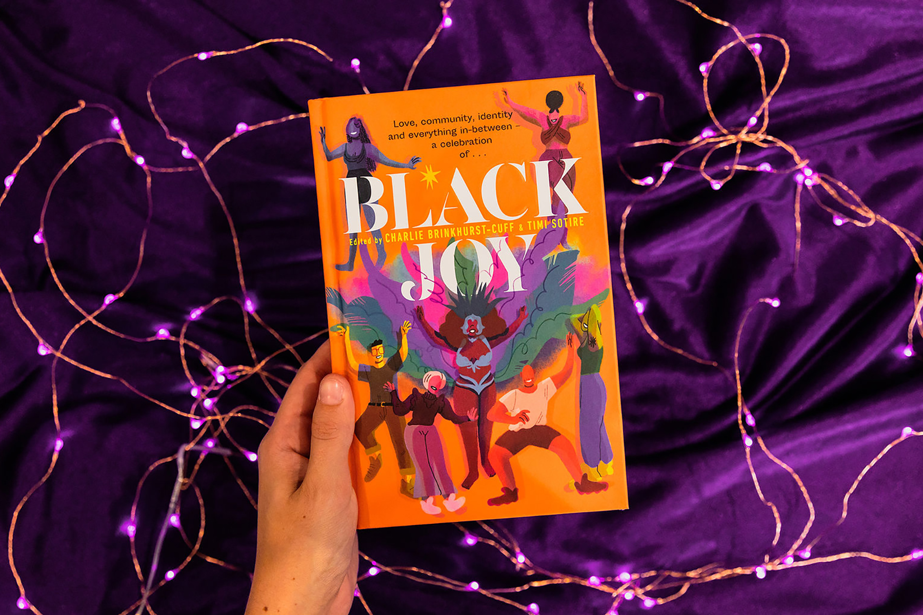An image of the book Black Joy being help by a hand in front of a purple material background with purple fairy lights