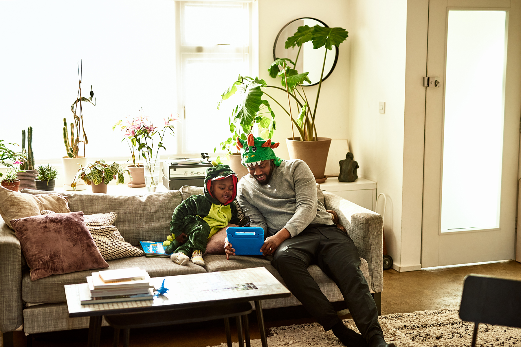 A photo of a dad and his son sitting on a sofa dressed as dinosaurs whilst they read a book together
