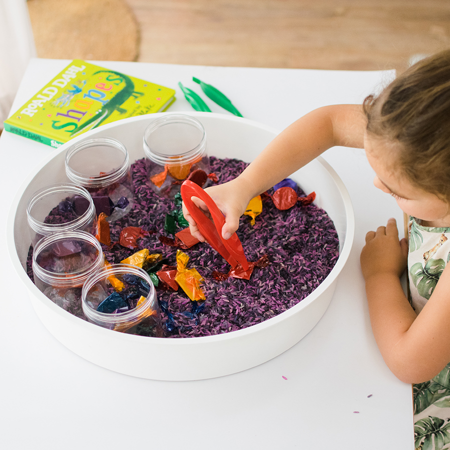 A photo of the Willy Wonka shape sorter being used by a young child. The little girl is holding a pair of plastic tweezers and picking out sweets of the bowl