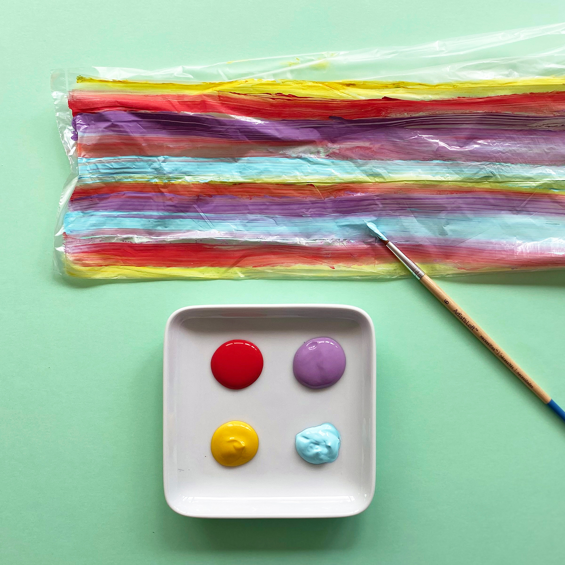 A photo of a piece of cellophane being painted in rainbow colours alongside a dish with paint and a paintbrush on a mint green background