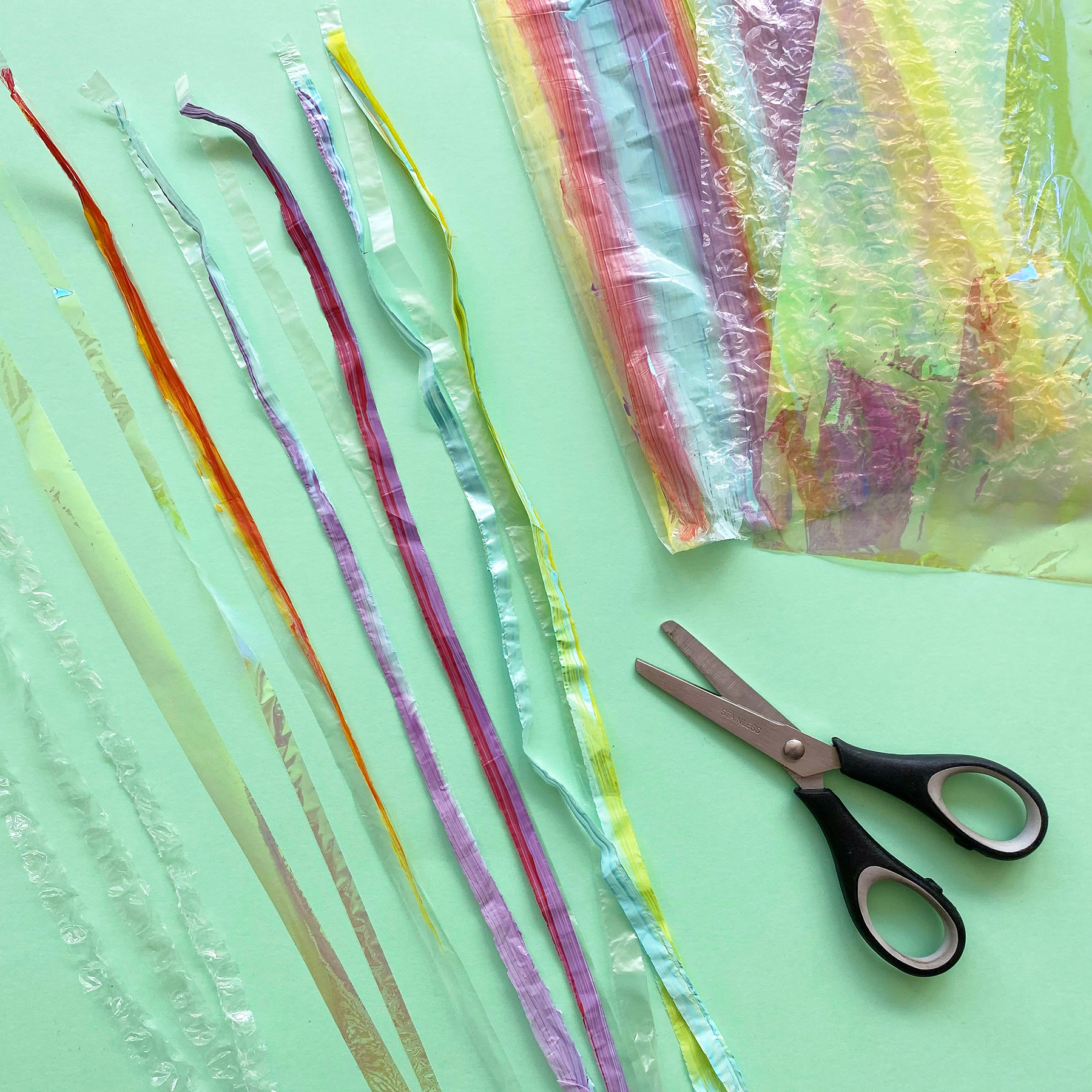 A photo of a pair of scissors alongside a painted plastic bag and cellophane that has been cut into long thin strips on a mint green background